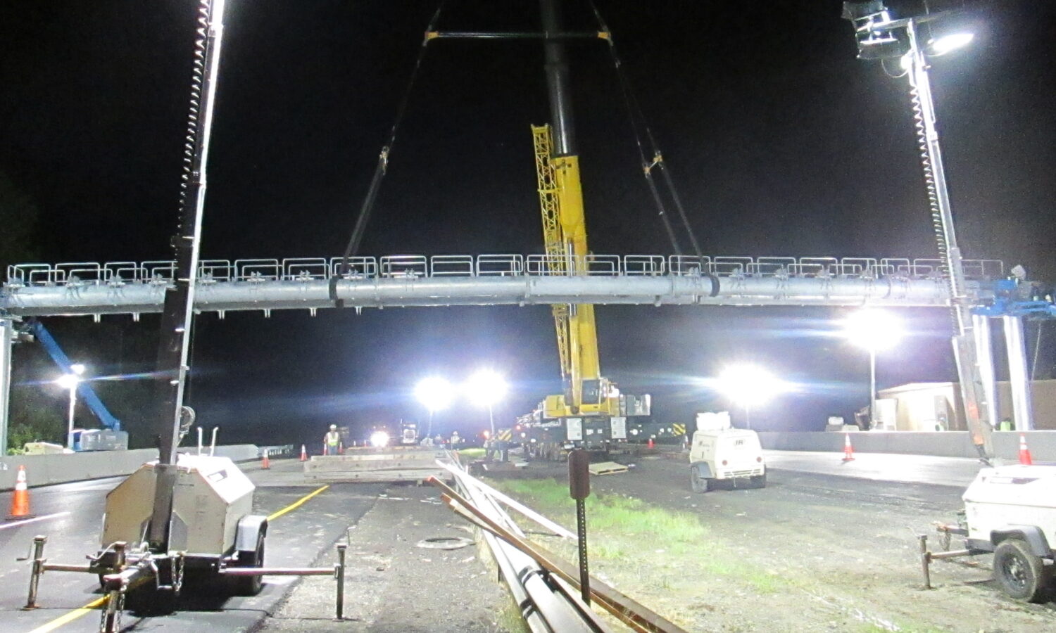 Crane lifts pedestrian bridge over road at night, lights shine.