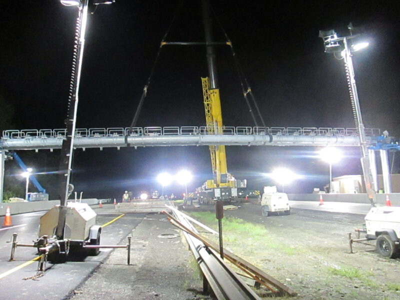 Crane lifts pedestrian bridge over road at night, lights shine.
