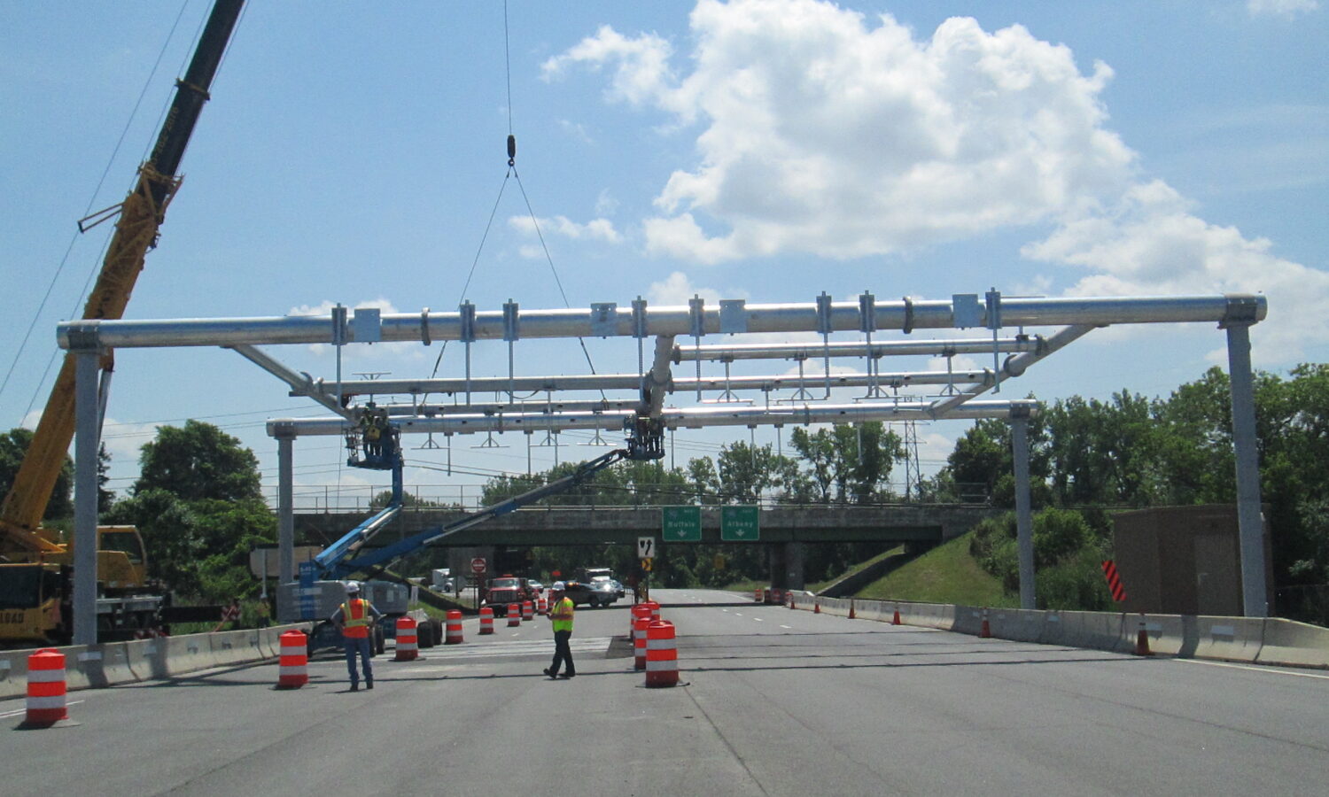 Workers guide overhead tolling gantry installed by crane; highway closed.