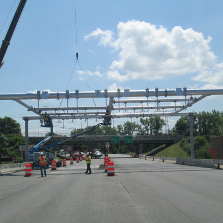 Workers guide overhead tolling gantry installed by crane; highway closed.
