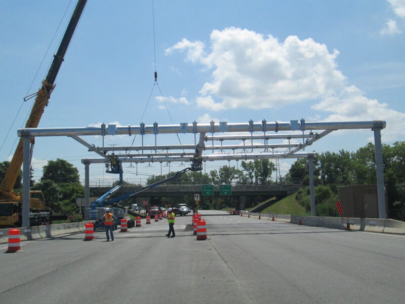 Workers guide overhead tolling gantry installed by crane; highway closed.