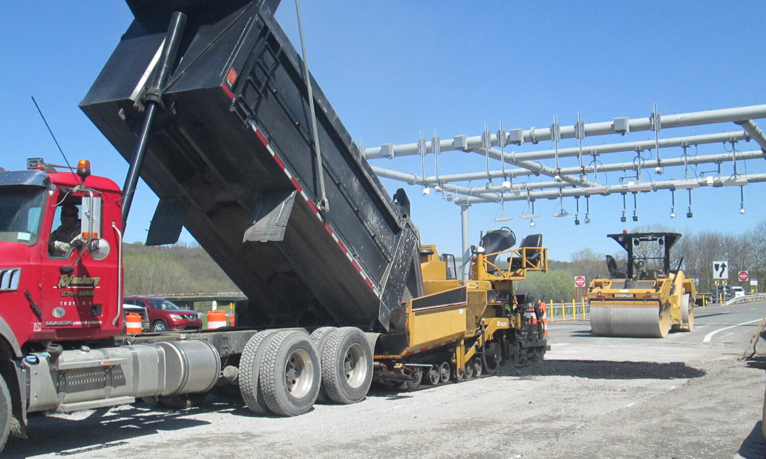 Dump truck unloads asphalt; roller compacts highway under clear sky.