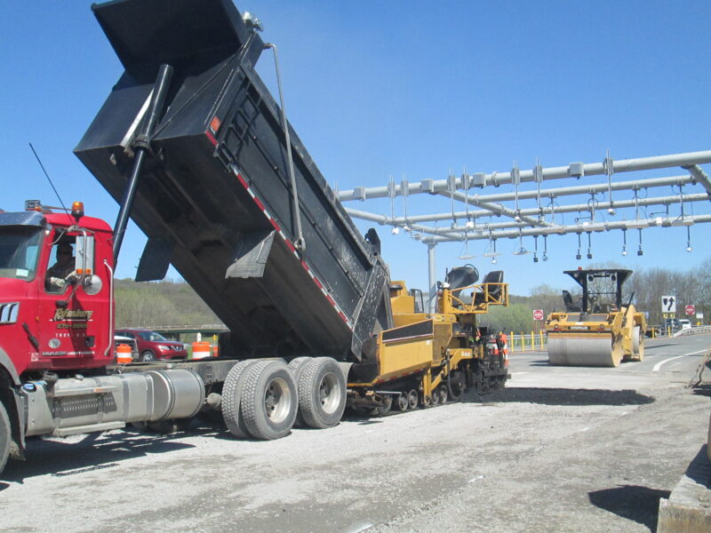 Dump truck unloads asphalt; roller compacts highway under clear sky.