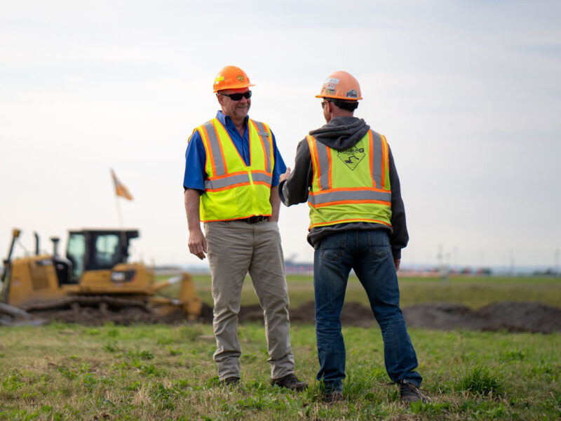 Two workers in safety gear shake hands in a grassy field