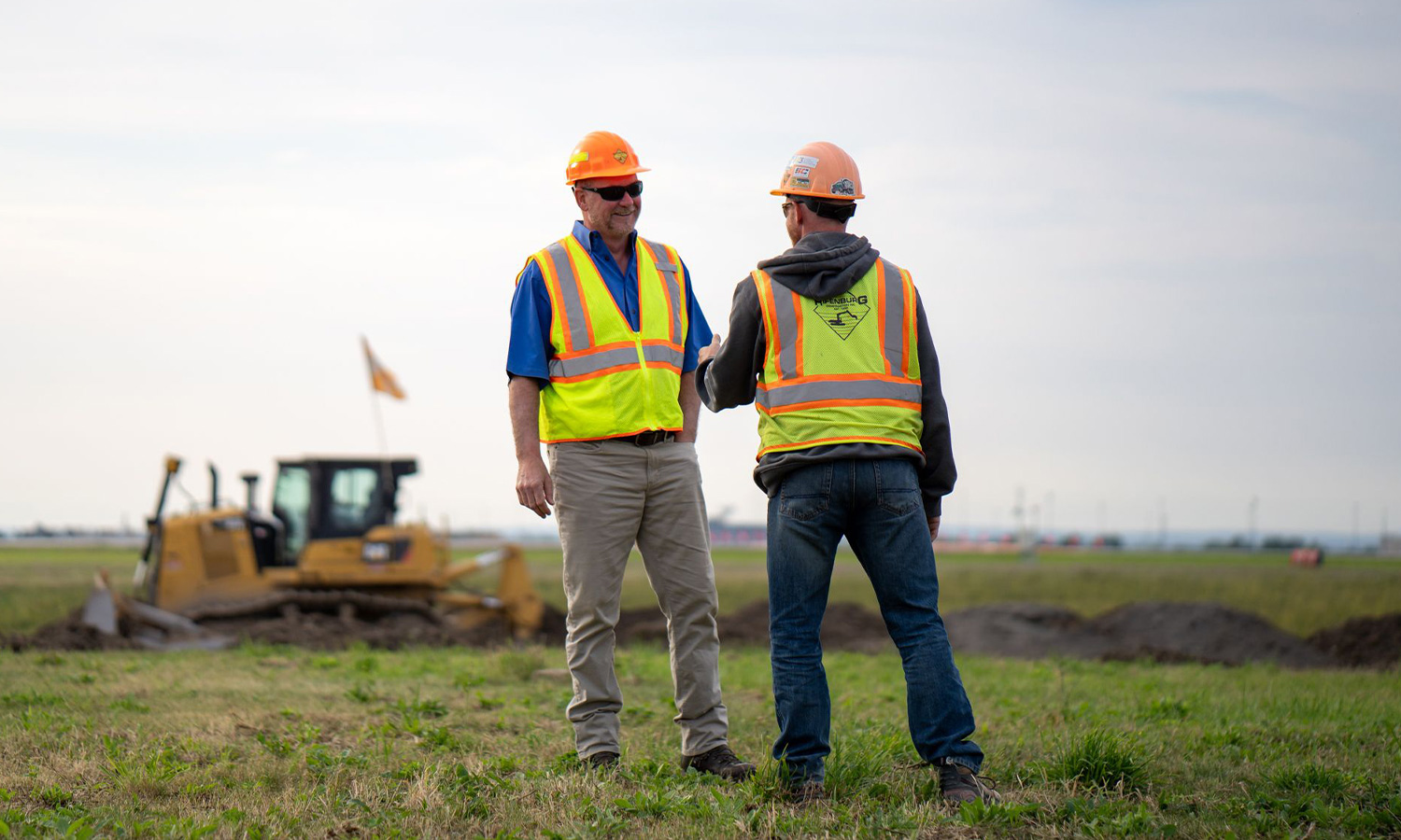 Two workers in safety gear shake hands in a grassy field