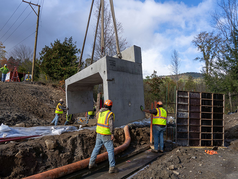 Workers guide concrete structure into trench using crane.