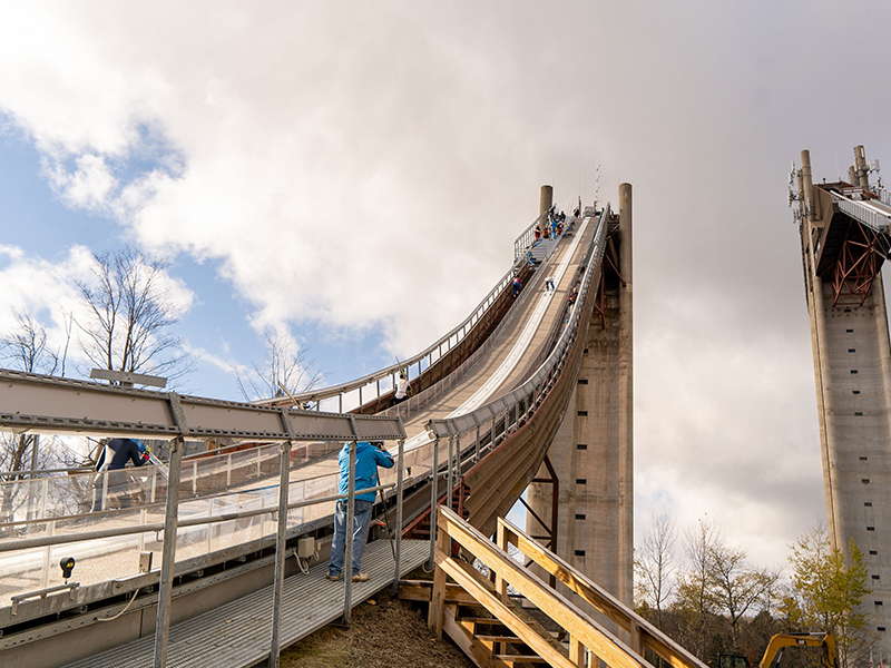 Outdoor ski jump with a skier descending and others at the top.