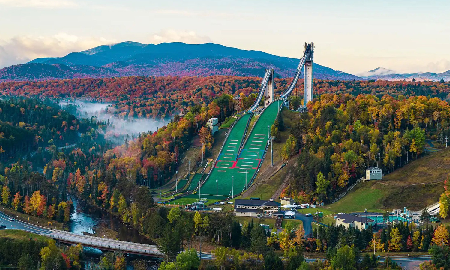 Large ski jump, autumn trees, mountains behind, river in front.